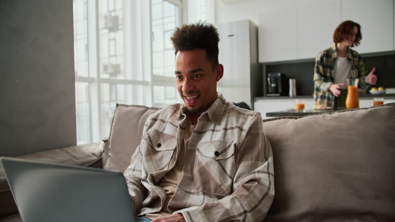 Happy Black-skinned brunette man with stubble in a checkered cream shirt works with his gray laptop and is very happy about his success while working from home in a modern studio apartment