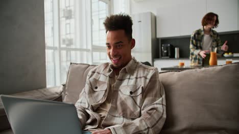 Happy-Black-skinned-brunette-man-with-stubble-in-a-checkered-cream-shirt-works-with-his-gray-laptop-and-is-very-happy-about-his-success-while-working-from-home-in-a-modern-studio-apartment