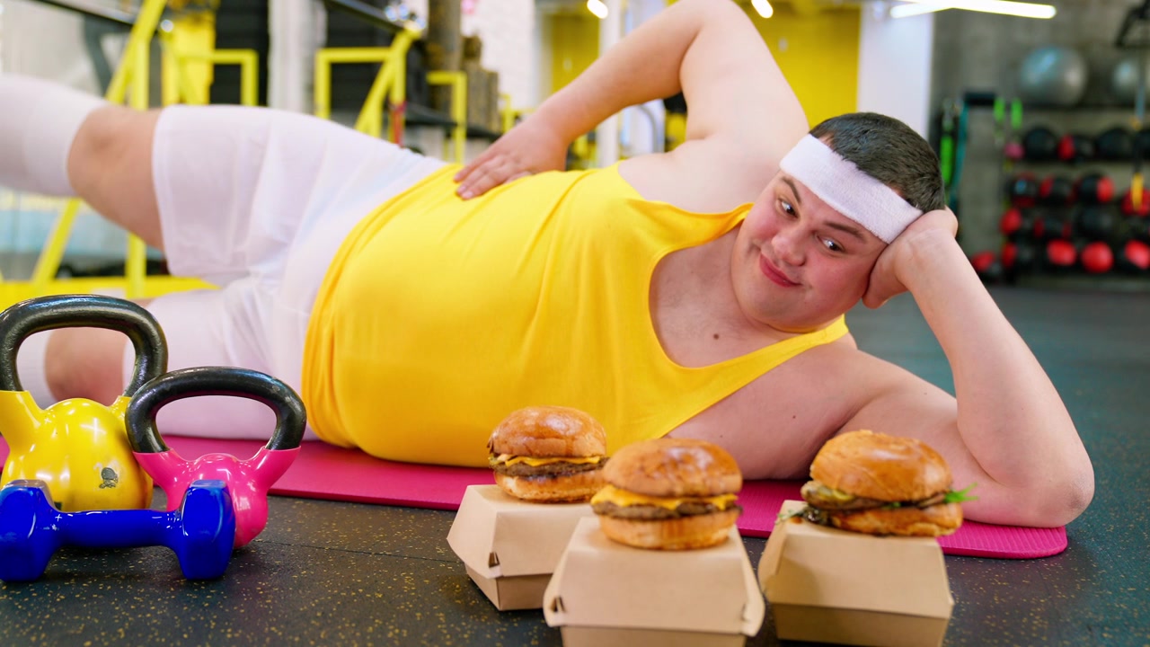 Uomo in sovrappeso su un tappetino da yoga che guarda gli hamburger in palestra