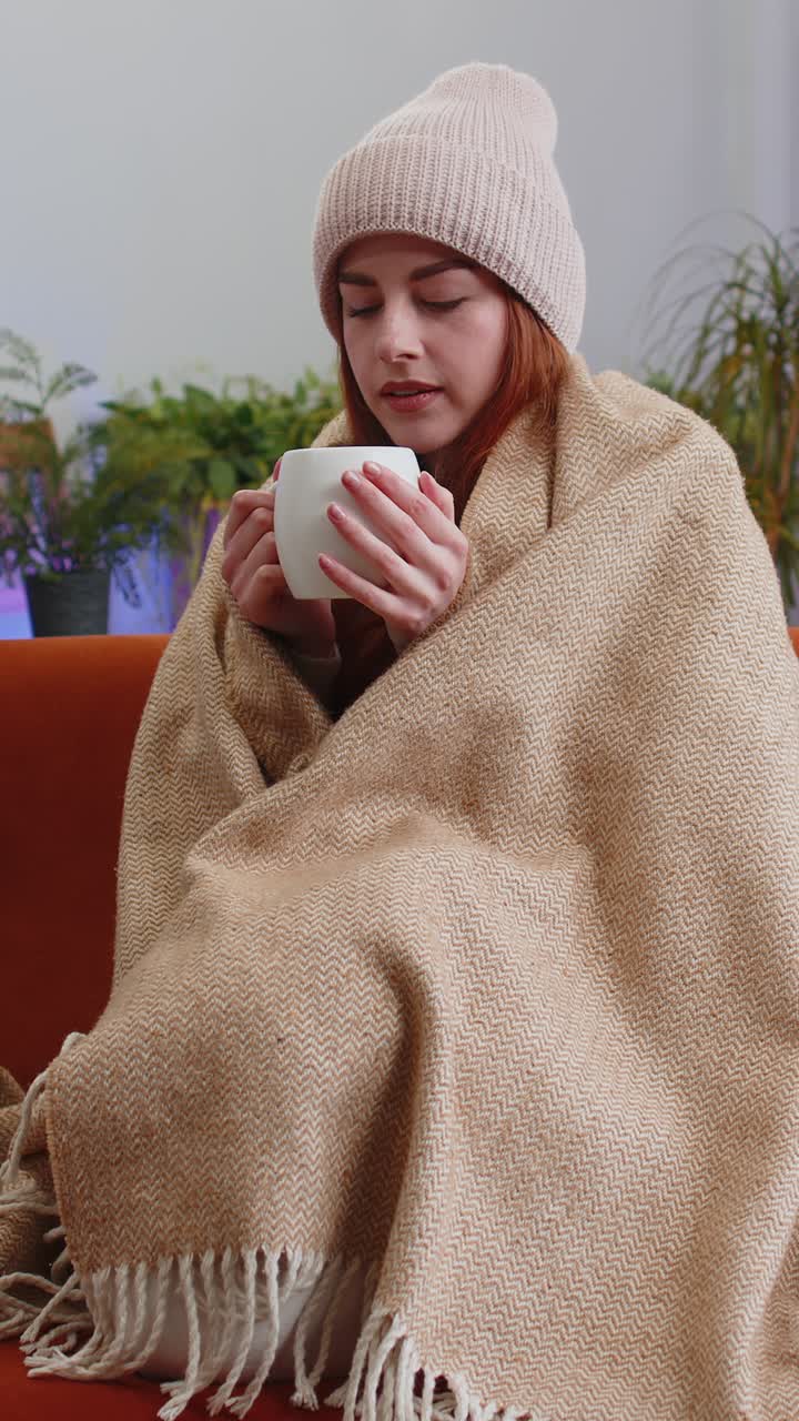 Una donna dai capelli rossi malata, con un cappello e avvolta in un plaid, siede da sola sul divano, tremando dal freddo e sorseggiando tè caldo.