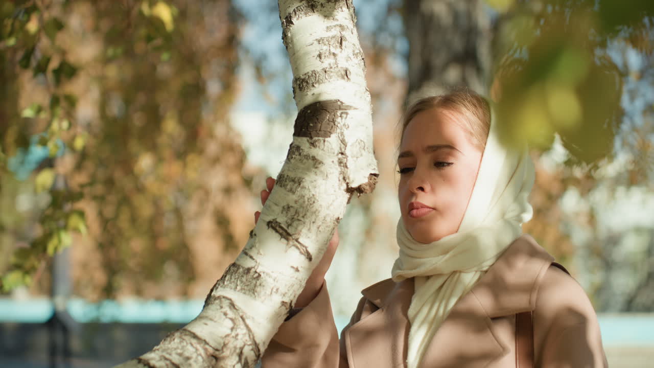 Giovane donna caucasica che abbraccia una betulla, parco autunnale, foulard color crema, cappotto beige, tocco delicato, occhi chiusi, calda luce solare, foglie che cadono, umore contemplativo, profondità di campo cinematografica ridotta.