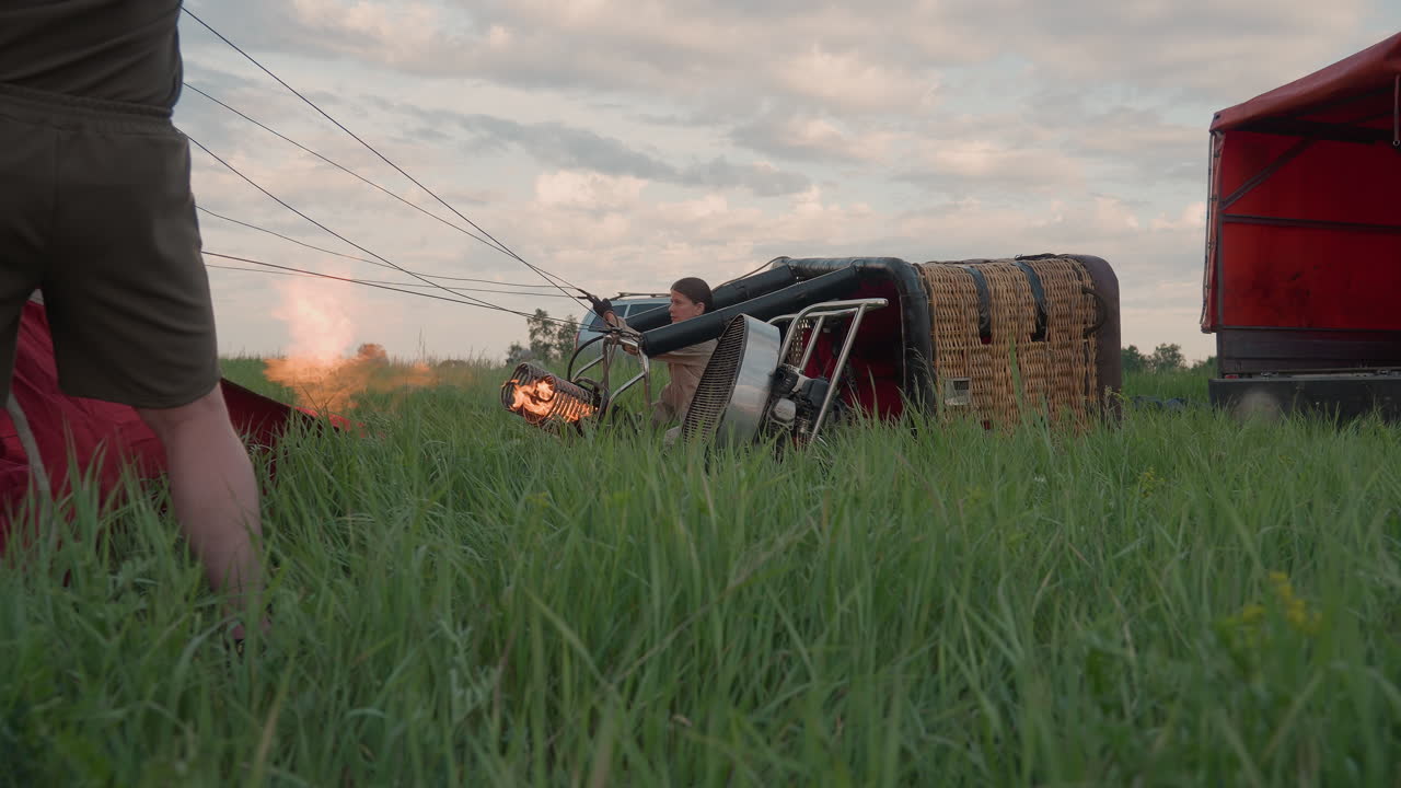 back view man in green shirt and shorts holds colorful hot air balloon envelope while woman crouches by wicker basket igniting burner flame in tall grass field under soft sunset sky