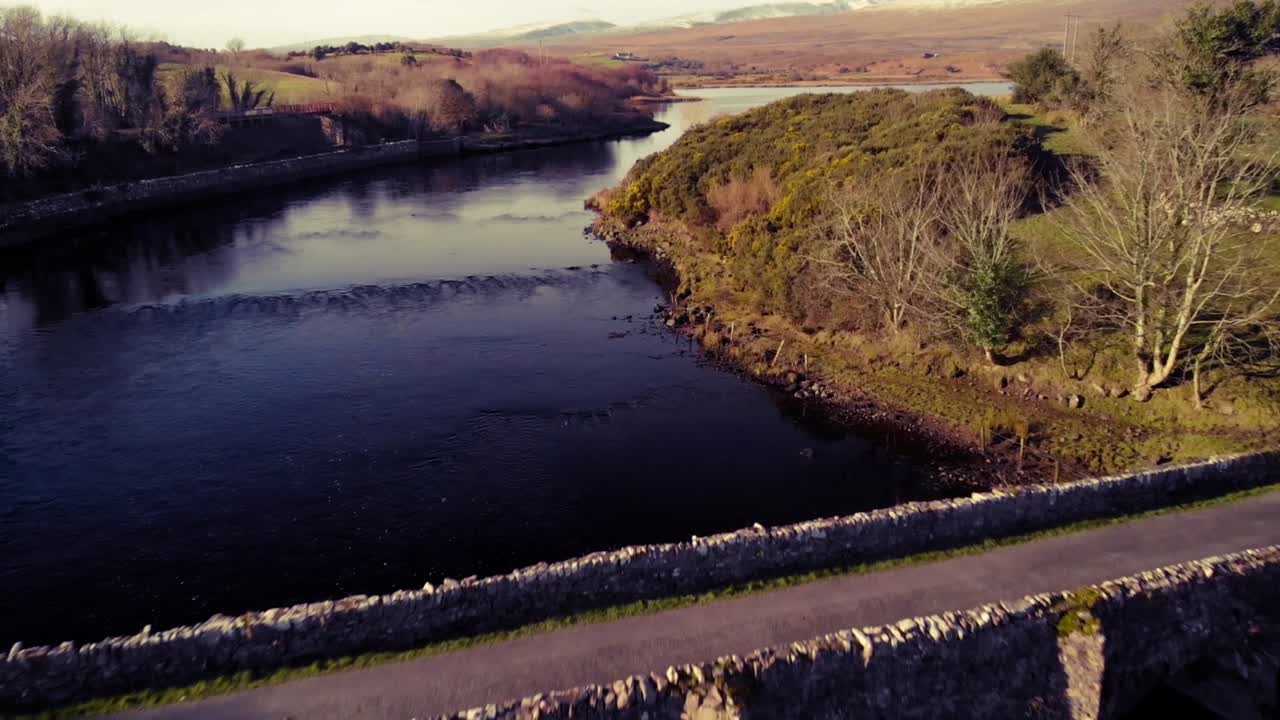 Flyover old stone bridge in Mayo, Ireland with snow capped mountains in the background. Blue sky and water, yellow grass on the banks