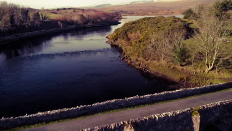flyover old stone bridge in mayo, ireland with snow capped mountains in the background. blue sky and water, yellow grass on the banks