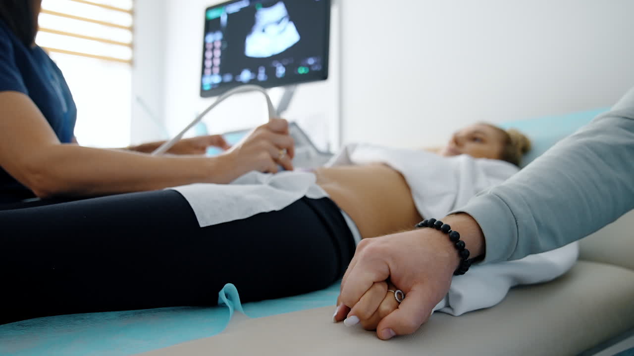 Male hand holding the hand of a lady lying on the couch. Pregnant woman having ultrasound examination in hospital. Blurred backdrop.
