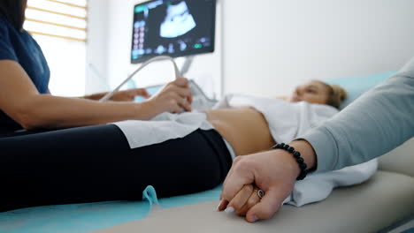male hand holding the hand of a lady lying on the couch. pregnant woman having ultrasound examination in hospital. blurred backdrop.