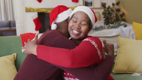 Glückliches-älteres-Afroamerikanisches-Paar-Mit-Weihnachtsmützen,-Das-Sich-Im-Wohnzimmer-Umarmt