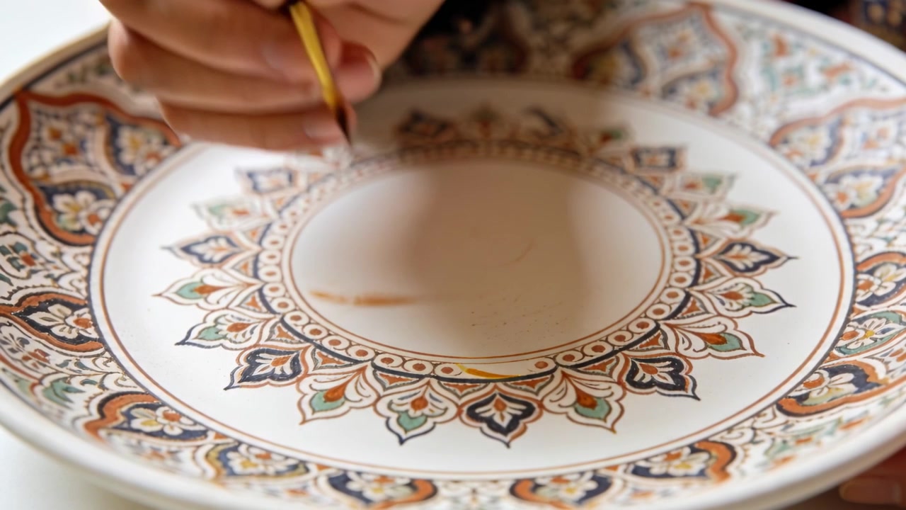 Mano destra decorata che tocca il bordo, applicando una linea dorata su un piatto di ceramica con un pennello, rifinendo.