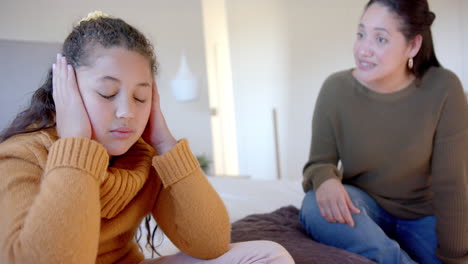 nervous biracial mother admonishing daughter on bed in sunny bedroom