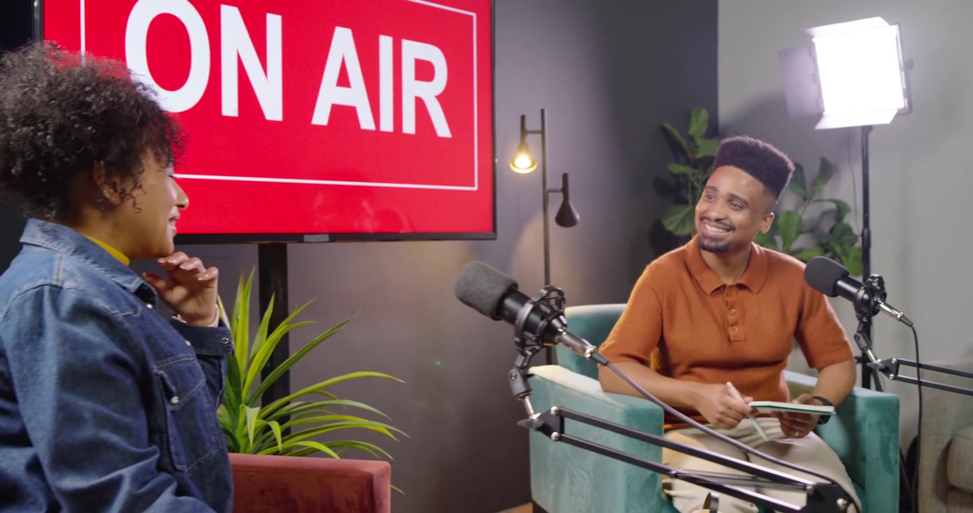 African American man host reacting to ON AIR cue, speaking into mics, taking notes, hosting guest