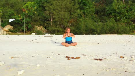 Schöne-Glückliche-Frau-Mit-Einem-Lächeln,-Die-Yoga-Und-Meditation-Am-Strand-Mit-Weißem-Sand-Und-Tropischer-Vegetation-Praktiziert