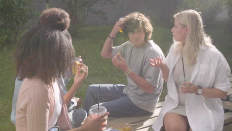 group of multiethnic friends spending time together, talking to each other, having fun and laughing while eating nuggets and drinking refreshing juice, sitting in a wooden bench in a park