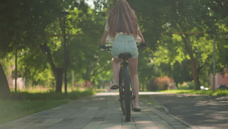 vista trasera de una mujer joven con confianza montando en bicicleta a través de un pintoresco sendero verde del parque, cabello largo que fluye naturalmente con el movimiento, la luz del sol se filtra a través de los árboles, proyectando sombras suaves