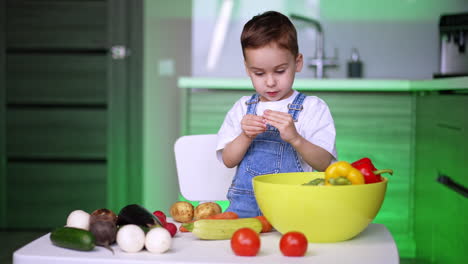 caucasian baby in white t-shirt and jeans romper stands at the table with multiple veggies. little kid learns vegetables at home.