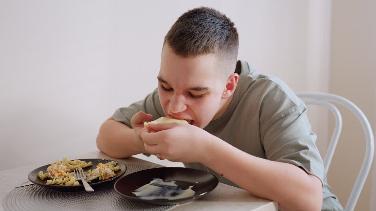 Boy in green shirt dips pancake into cream and eats slowly at dining table, two plates in front of him including scrambled pasta, peaceful indoor mealtime