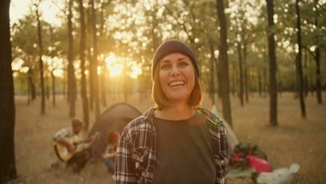 a happy middle-aged girl in a hat and bob hairstyle smiles and grimaces at the camera against the backdrop of a green, sunny summer forest and the rest of the hiking group
