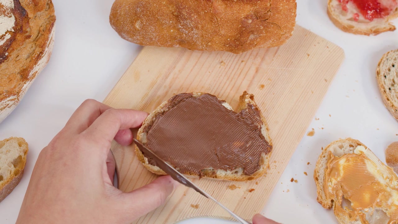 Top view of hands spreading chocolate cream on bread over a wooden board.