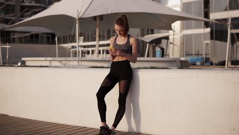 slim, attractive girl in sports clothes looking at mobile phone leaning on a white parapet. seaside. relaxing, smiling. enjoying her time. sun shines