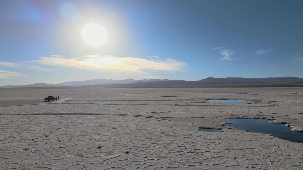 Off road vehicle drives across the cracked white surface of Salinas Grandes salt flat with bright sun and distant mountains, showing a wide desert landscape and sense of isolation.
