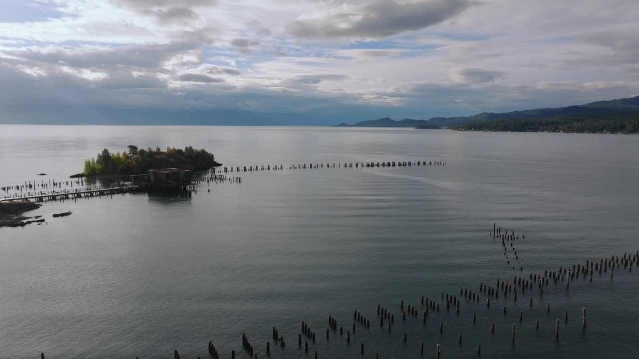 Tiang tambat dermaga panjang membentang melintasi air danau yang memantulkan cahaya dengan garis pantai yang jauh dan pantulan awan lembut di permukaan yang tenang, Danau Flathead Montana AS.