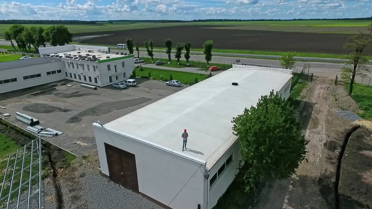 Male worker standing on a roof on the nature background. Engineer in protective helmet on a building among the field with future solar power station. Camera rising up.
