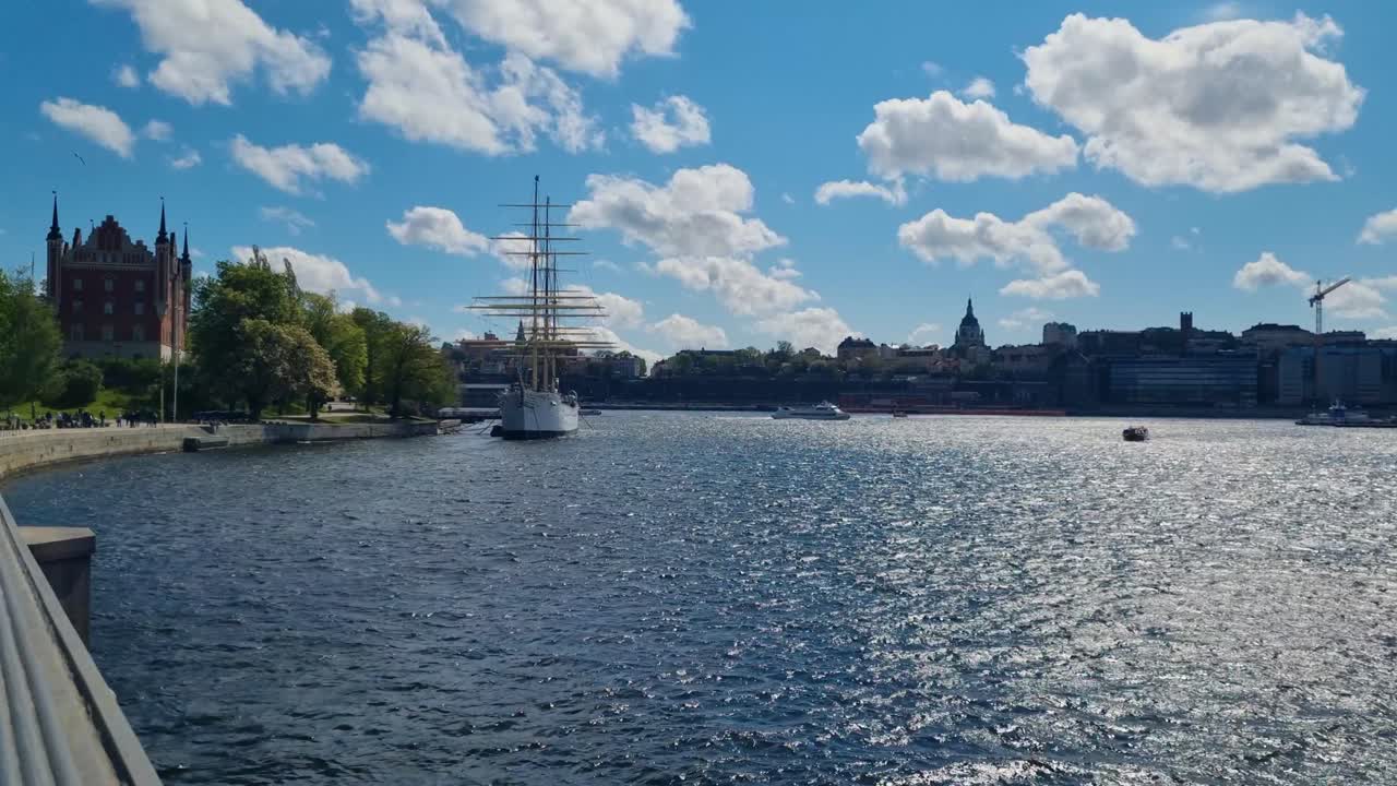 Panoramisk udsigt over Saltsjön bugten i Stockholm med det fuldt riggede sejlskib af Chapman, der ligger fortøjet ved Skeppsholmen, under en klar blå himmel og et roligt nordisk lys.