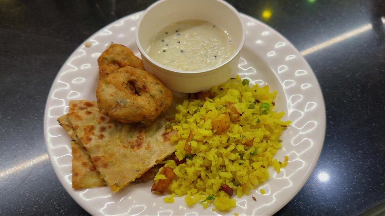Cámara orbitando alrededor de un plato de desayuno tradicional indio que incluye poha, medu vada, chutney de coco y paratha doblado, dispuestos en una fuente blanca.