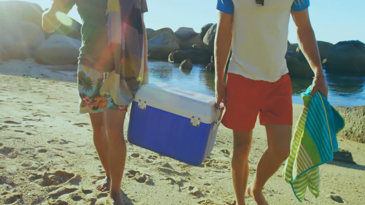 Carrying cooler and towels, two people walking on sandy beach near water
