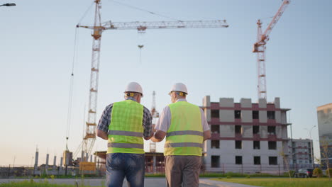 waist-up of two middle-aged male builders wearing safety clothing standing at construction site man using walkie-talkie his colleague holding paper with project plan