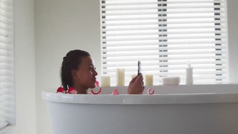 african american woman using smartphone in the bath tub in the bathroom at home