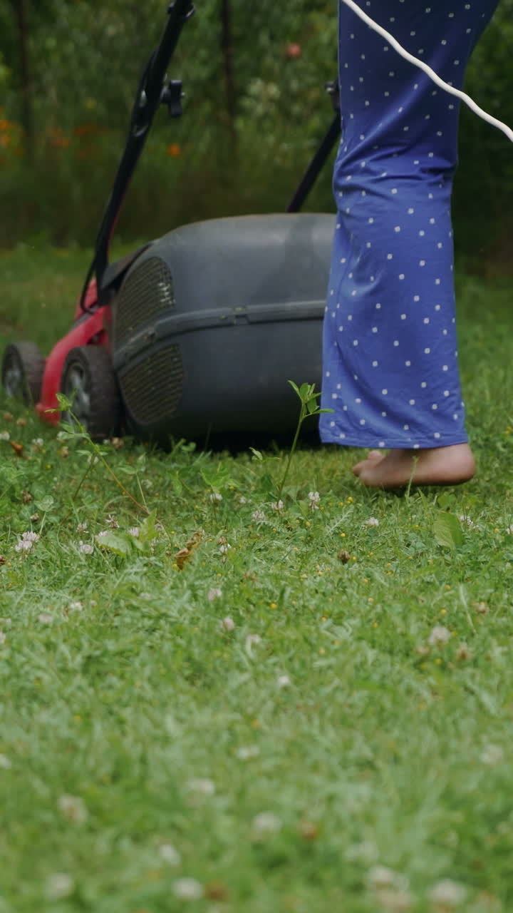 human is controlling the work of lawn mower near the path in the yard in the summer. The lawn mower consistently is mowing grass Vertical video