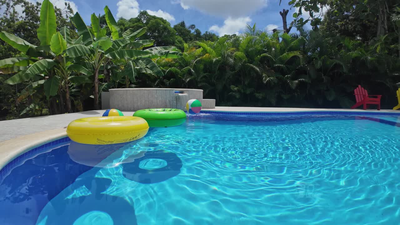 Tropical swimming pool with colorful floats, waterfall, and lush greenery in the background. Establishing static shot