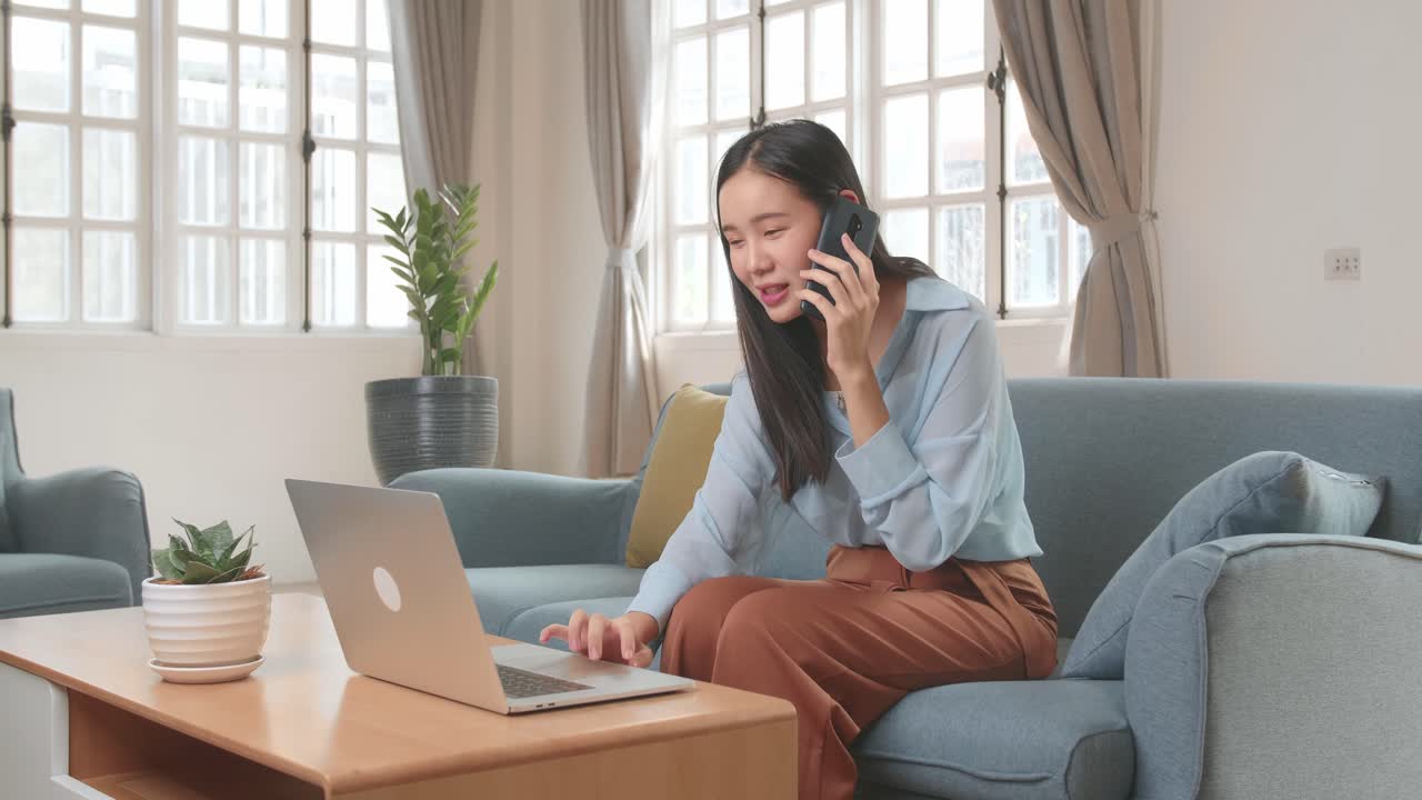 リビングでソファに座り、電話で話しながらノートPCで作業するアジア人女性、4Kビデオ