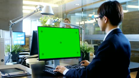 in the office businessman works on a desktop personal computer with mock-up green screen. colleague enters office and takes place at her desk.