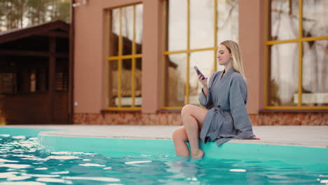 Una-Mujer-Bonita-Está-Sentada-En-El-Borde-De-Una-Piscina-Termal-Al-Aire-Libre-Y-Descansando-Navegando-Por-Internet-Con-Un-Teléfono-Inteligente-Moderno