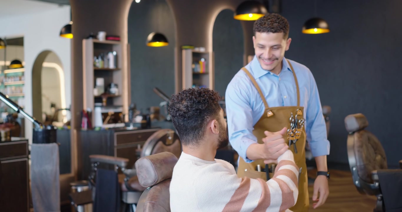 To afrikanske mænd; kunde scroller på telefon, frisør går i forklæde, hilser og styler hår i salon.