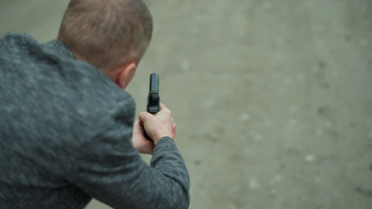 una vista de cerca desde atrás de un hombre en una chaqueta gris apuntando una pistola a un objetivo invisible por un camino desierto