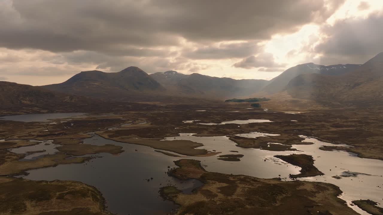 Aerial drone footage of the dramatic Glencoe valley in the Scottish Highlands, captured in moody pre-evening light. Sweeping views of rugged mountains, winding roads, and wild, untouched nature