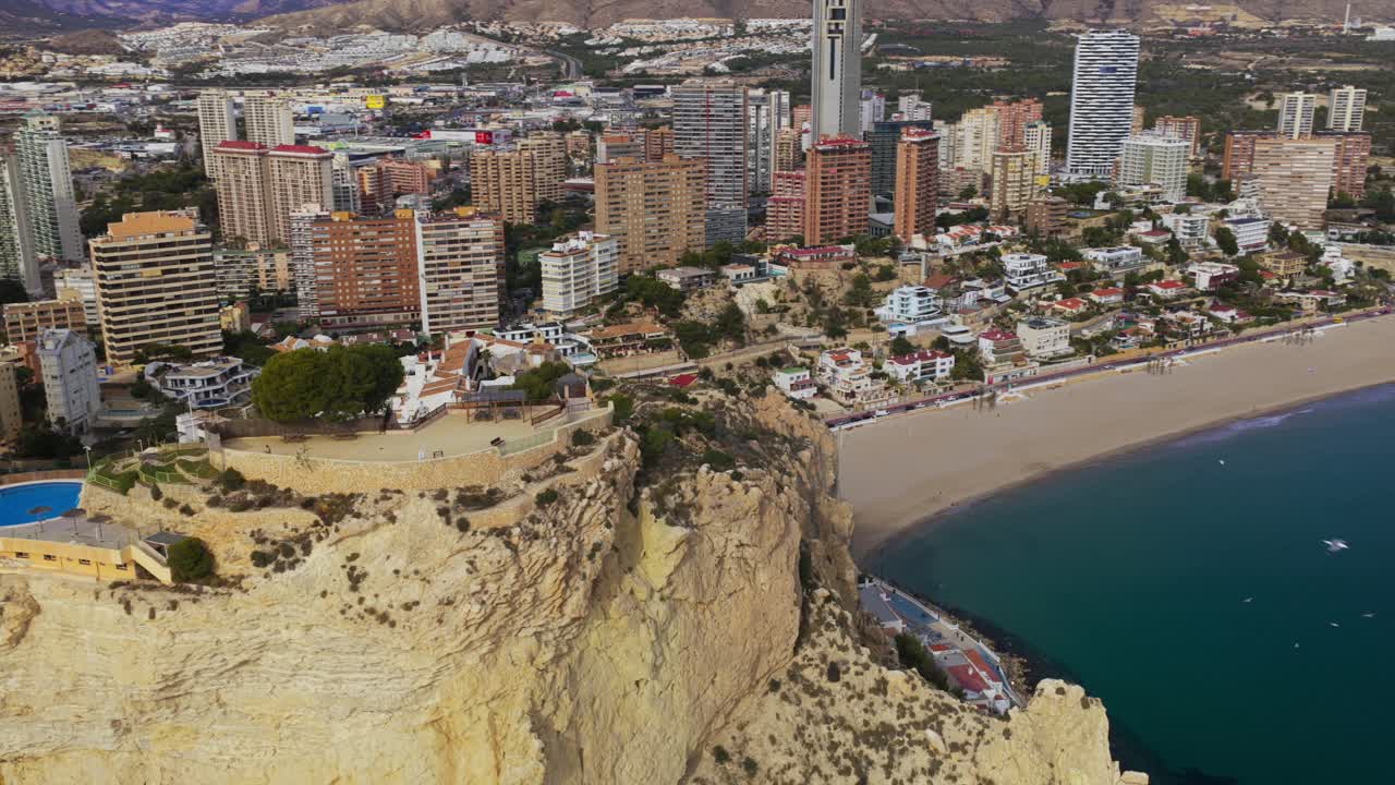 Luftfoto af Benidorm, Spanien, der viser bybilledet og kystlinjen. En sandstrand, turkist hav og moderne arkitektur under en lys himmel, hvilket gør det til en populær rejsedestination.