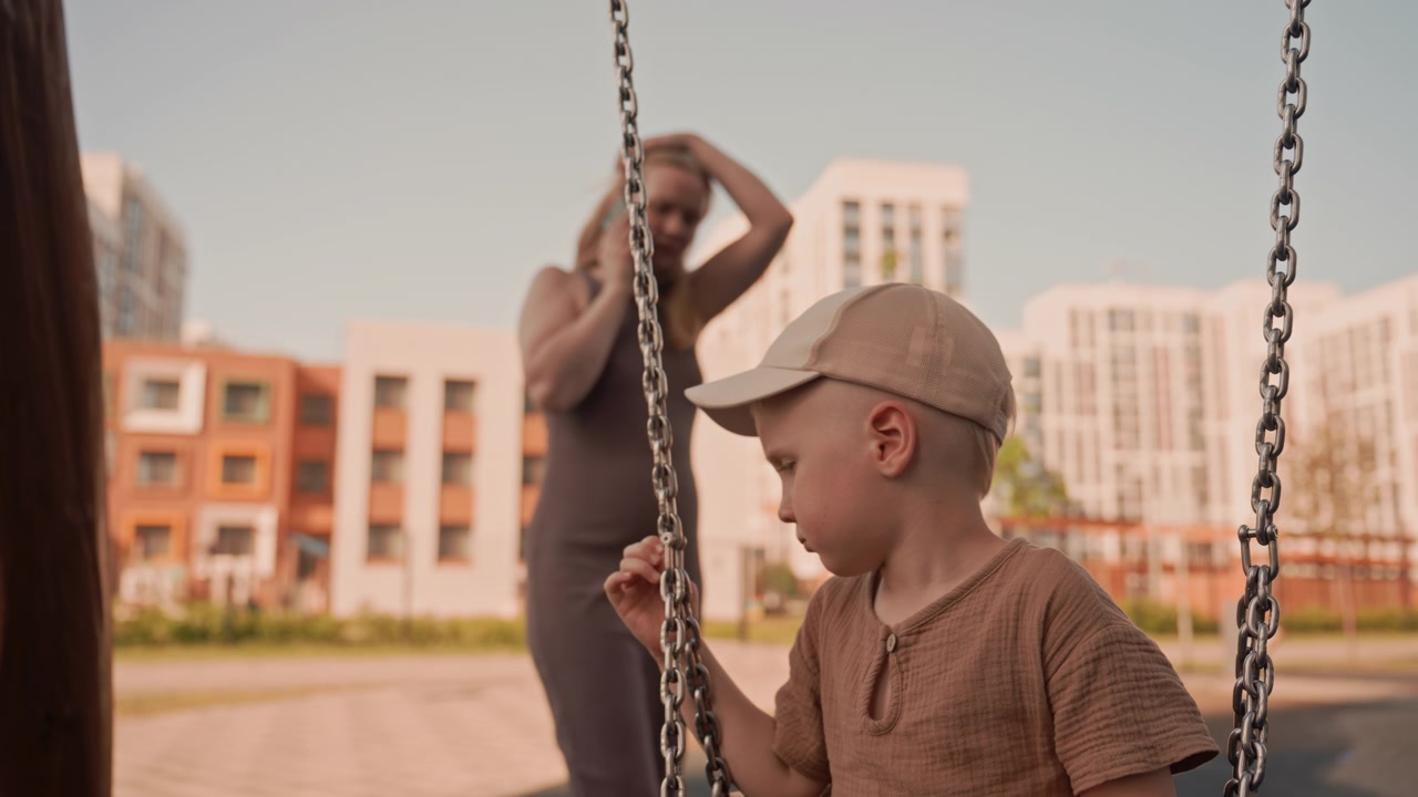 Un bambino siede tranquillamente su un'altalena in un parco giochi all'aperto, indossando una camicia marrone e un berretto. Una donna adulta è in piedi nelle vicinanze, parzialmente visibile, mentre il bambino si tiene alla catena. Lo sfondo presenta edifici moderni.