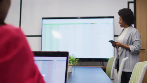 African-american-businesswoman-using-tablet-presenting-at-screen-to-female-colleague-at-meeting