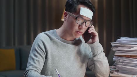 close up of sick asian man talking on phone and taking note while working hard with documents at home