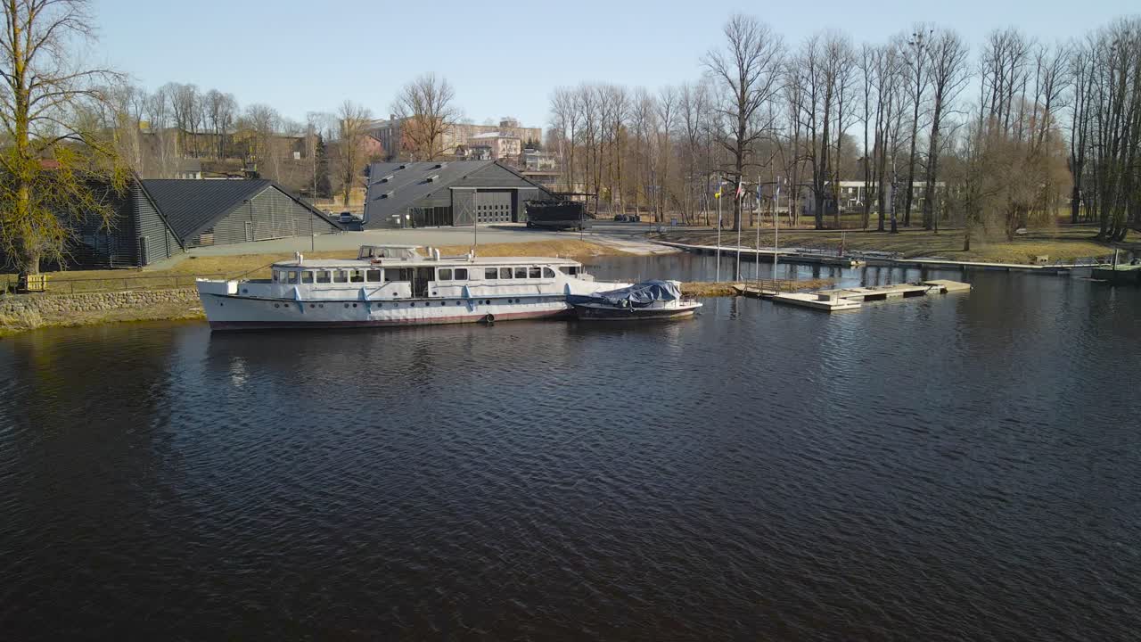 Aerial drone footage orbiting and circling around a white metallic boat that is near a wooden small harbor on Emajõgi or mothers river in Estonia Tartu during a beautiful sunny day. Water waves moving