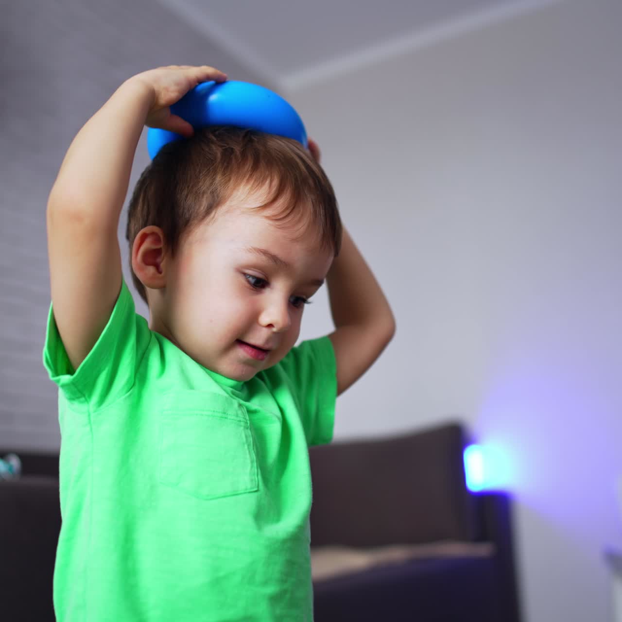 Cute toddler in light blue t-shirt puts the ring from a toy pyramid on his head. Happy kid having fun at home. Blurred backdrop