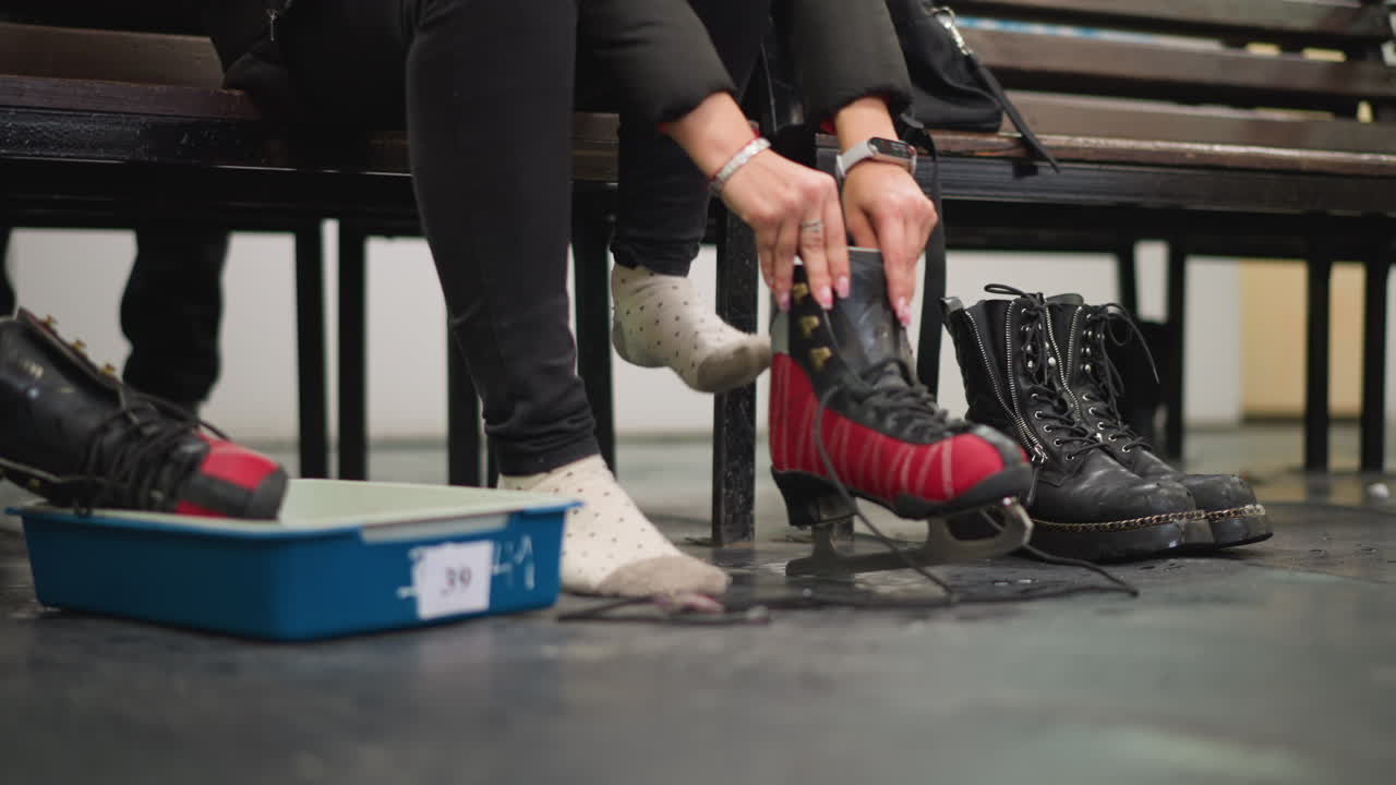 Female in polka dot socks sitting on bench putting on red black ice skates rest inside blue tray on wet locker room floor during footwear change after skating activity