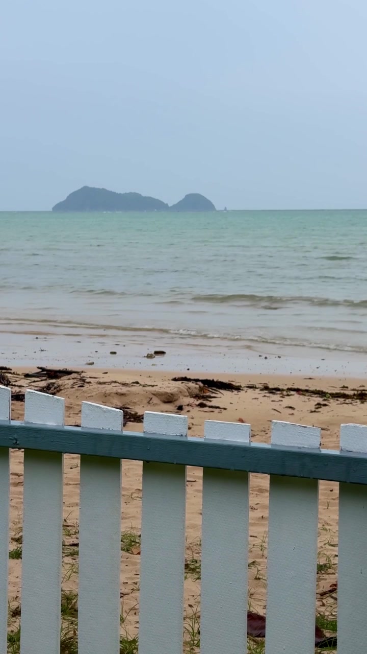 Rolig sandstrand med milde bølger og en fjern øy, sett over et hvitt stakittgjerde. Vertikalt reisevlog-opptak som fanger en fredelig ferie ved sjøen. feriestemning reise thailand