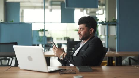 Porträt-Eines-Gutaussehenden,-Stilvollen-Jungen-Mannes-Arabischer-Herkunft,-Der-Mit-Einem-Laptop-In-Einem-Modernen-Businesscenter-Büro-Sitzt-Und-Auf-Seinem-Smartphone-In-Sozialen-Medien-Und-Messengern-SMS-Schreibt