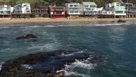 Beautiful-aerial-drone-shot-over-seals-relaxing-over-rocks-in-the-sea-leading-upto-the-luxurious-beachside-bungalows-in-Malibu,-California,-USA