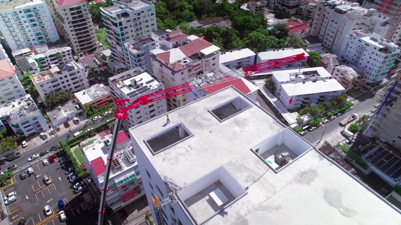Toma aérea de dron en picado, avanzando y ascendiendo sobre el tejado de una obra en construcción con una grúa telescópica roja, revelando el paisaje urbano circundante en Santo Domingo