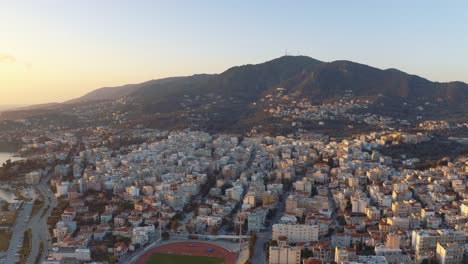 Sunrise-Aerial-drone-Flight-over-mountains-to-downtown-mitilini-revealing-sea-with-ferry-to-athens,-lesvos,-greece